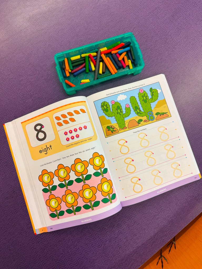 Open activity book showing the number '8' with illustrations of flowers and cacti, along with a tray of colorful crayons.
