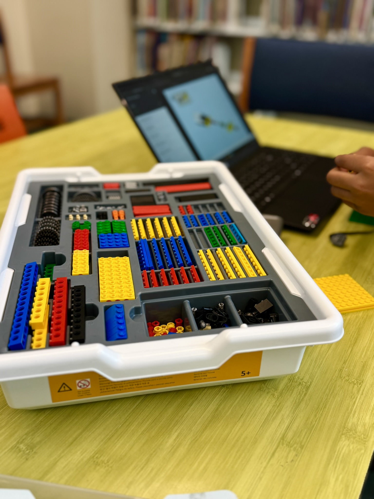 Tray with colorful LEGO pieces on a table with a laptop in the background and a person's hand reaching out
