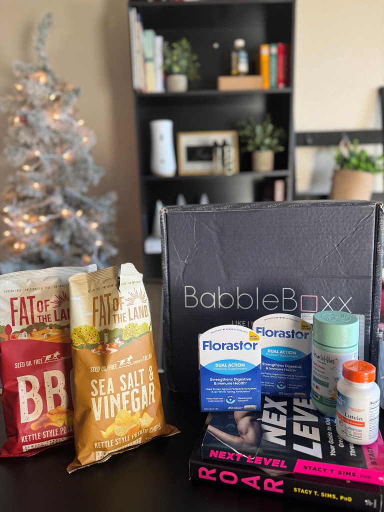 A display of winter wellness products including 'Fat of the Land' chips, 'Florastor' probiotic supplements, 'True Littles' multivitamin, and books by Dr. Stacy Sims laid out on a table with a Christmas tree in the background.
