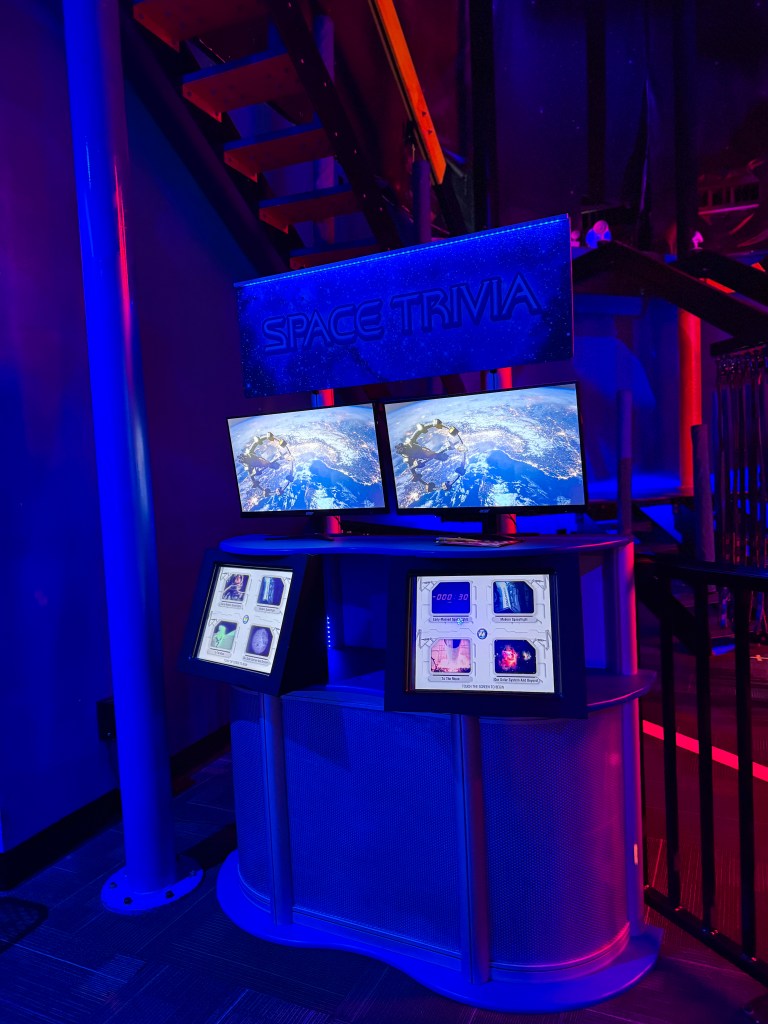 A space-themed trivia game station featuring two monitors displaying Earth and space images, accompanied by interactive touchscreens for game controls.