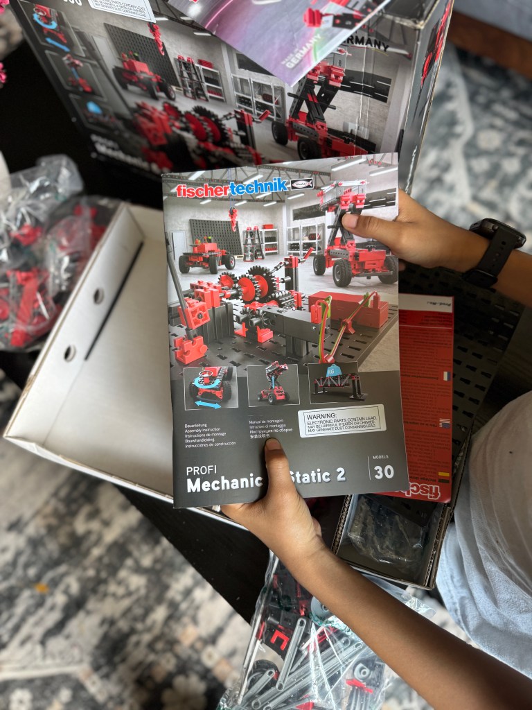 A child holding the Fischer Technik STEM kit instruction manual, surrounded by building pieces in a box. The manual features colorful graphics of mechanical models with a warning label.