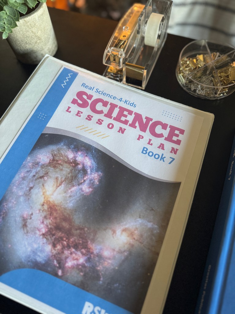Close-up of the 'Real Science 4 Kids Book 7' lesson plan binder featuring a colorful cover with a cosmic image, resting on a desk next to tape and paper clips.