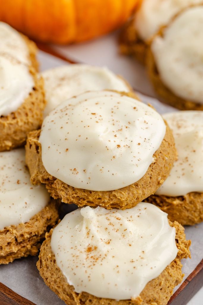 A close-up of soft pumpkin cookies topped with a creamy white frosting and a sprinkle of cinnamon, with a small pumpkin in the background.