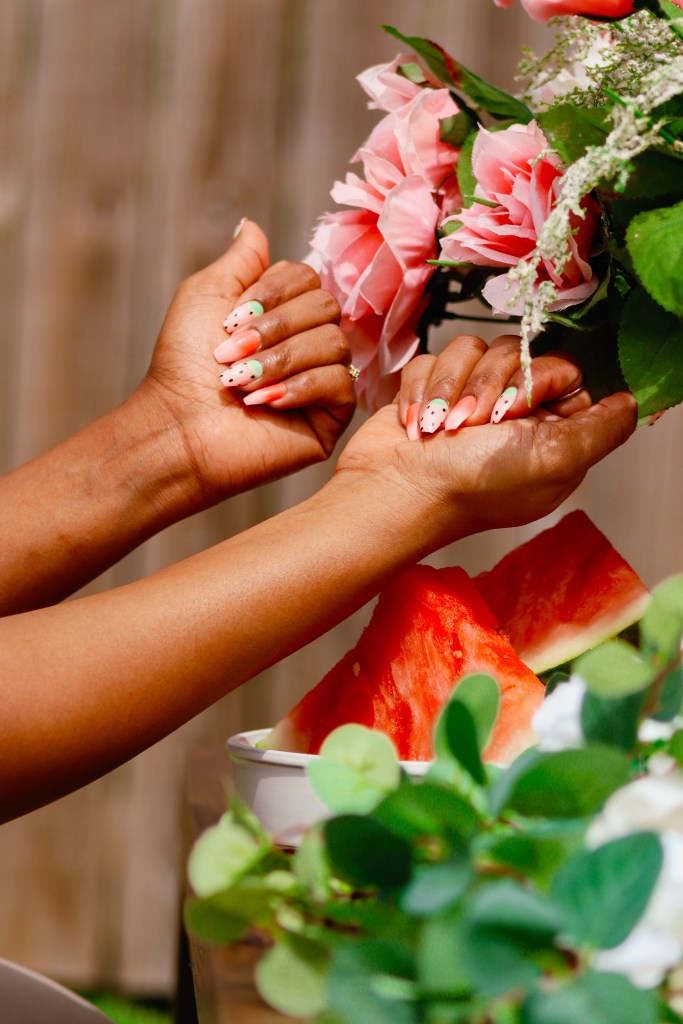 Close-up of two hands with colorful nail art surrounded by pink flowers and a bowl of watermelon slices.