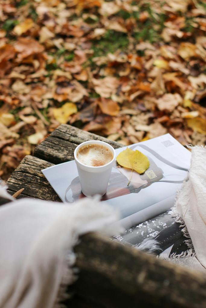 A cozy autumn scene featuring a cup of coffee placed on a magazine, surrounded by fallen leaves and a soft blanket. The warm colors evoke a seasonal vibe.
