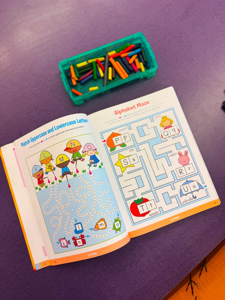 An open educational workbook featuring an alphabet maze and a matching uppercase and lowercase letters activity, with a container of colorful crayons on a purple table.
