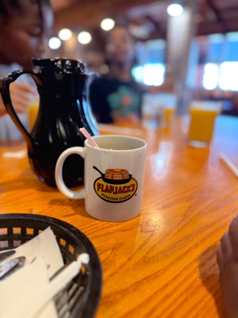 Flapjack's Pancake Cabin coffee mug on a wooden breakfast table with family members seated in the background.