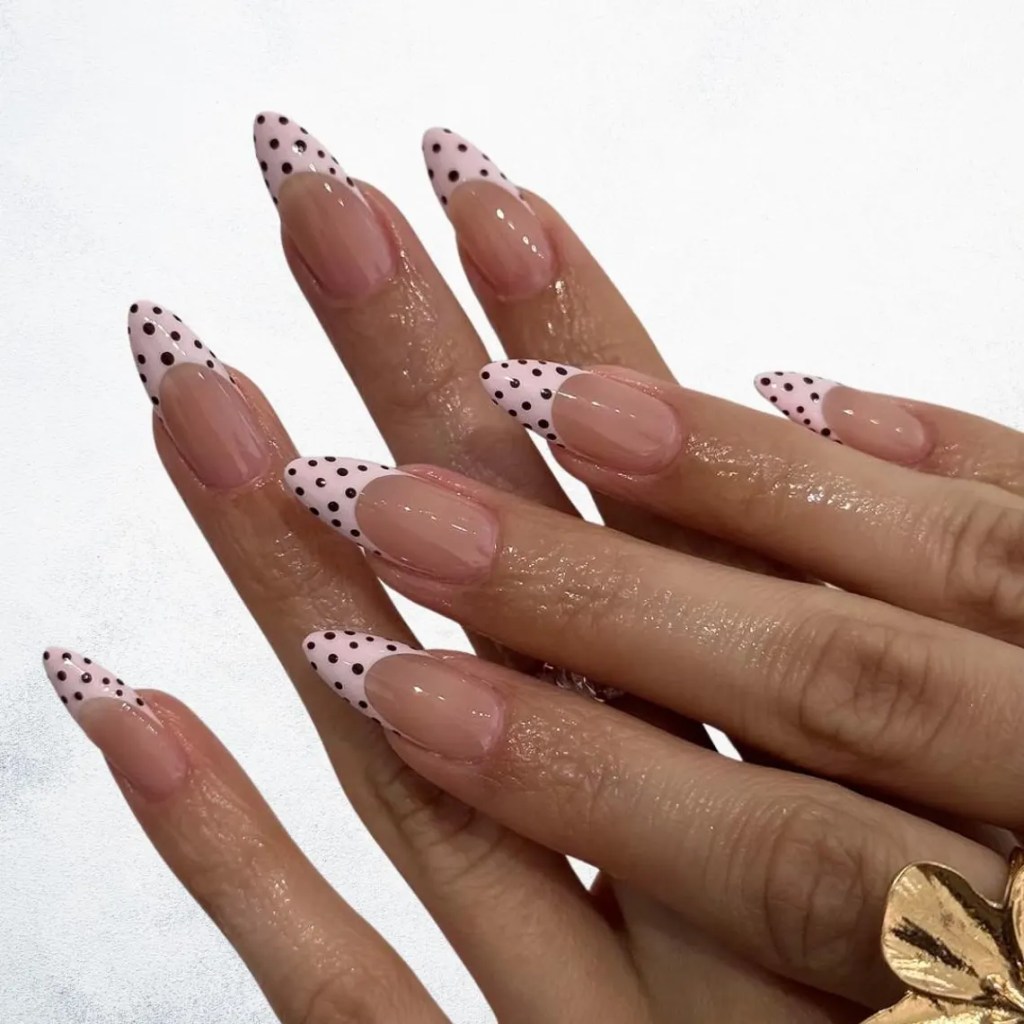 A close-up of hands with long almond-shaped nails featuring a nude base and pink tips adorned with black polka dots.