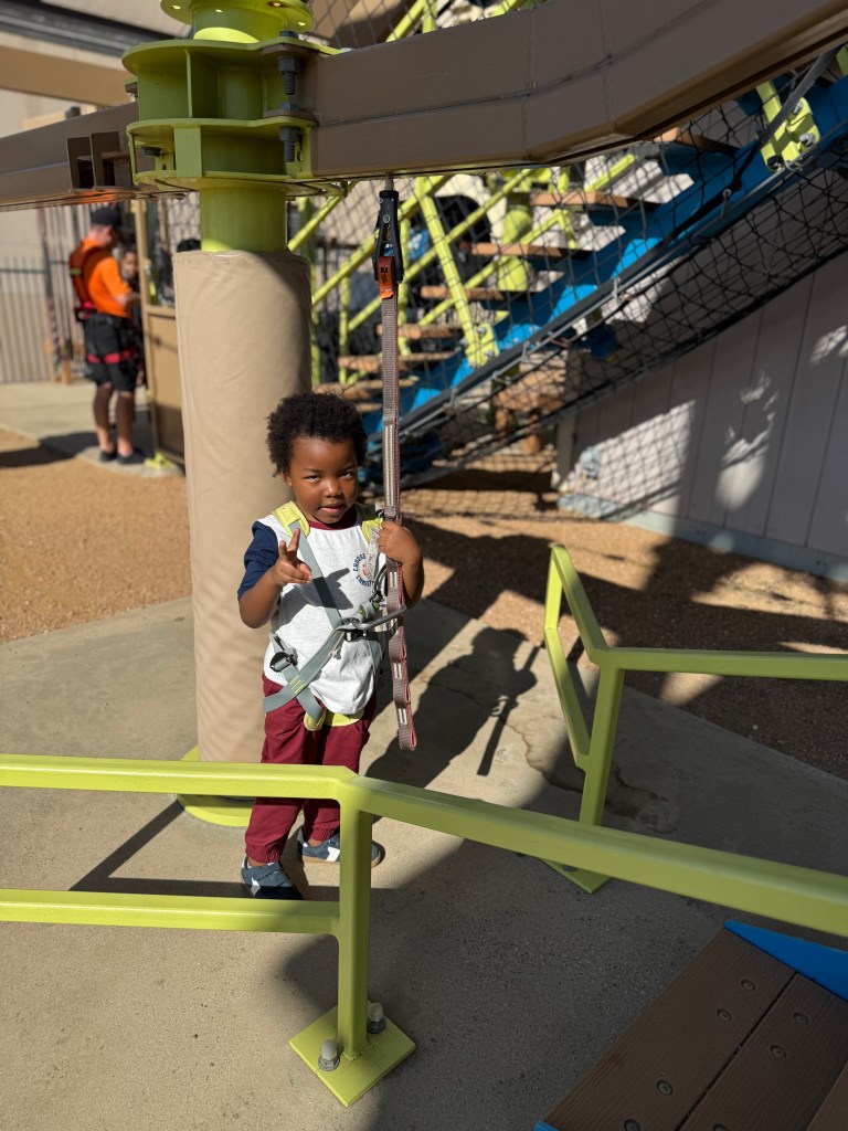 A young child in a harness, standing at the base of a ropes course, smiles and gives a thumbs up while holding onto the safety line.