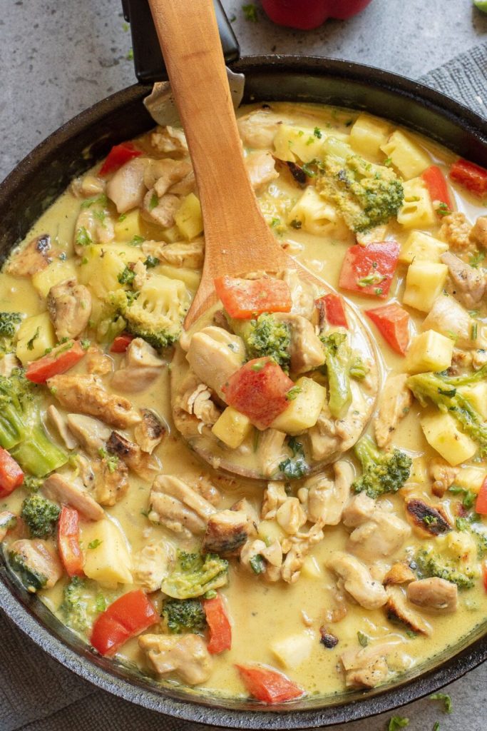 A close-up of a creamy chicken and vegetable stir-fry in a skillet, featuring chunks of chicken, broccoli, red bell peppers, and pineapple, with a wooden spoon stirring the colorful ingredients.