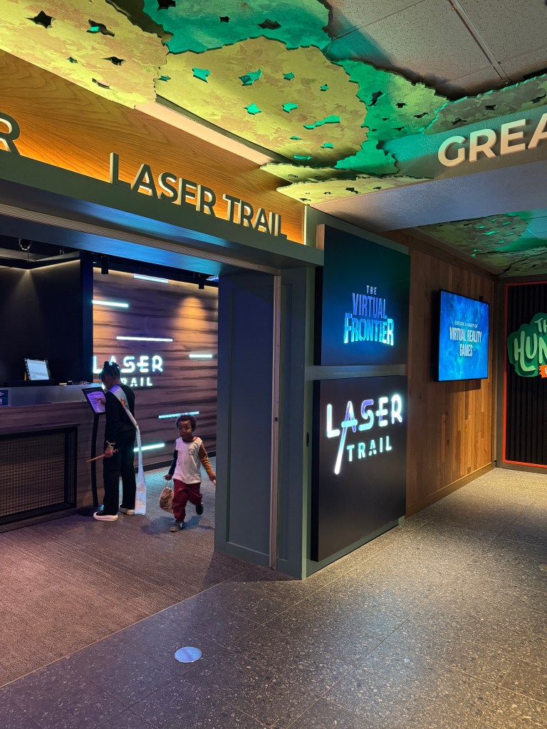 Entrance to the Laser Trail attraction at Great Wolf Lodge, featuring a sign with 'LASER TRAIL' and a colorful ceiling design. A child walks past, while an adult stands at the check-in area.