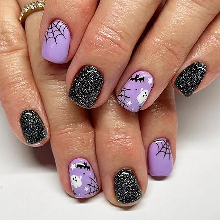 A close-up of hands showing Halloween-themed nails, featuring pastel lavender polish with cute ghost and spiderweb designs, along with black glittery accents.