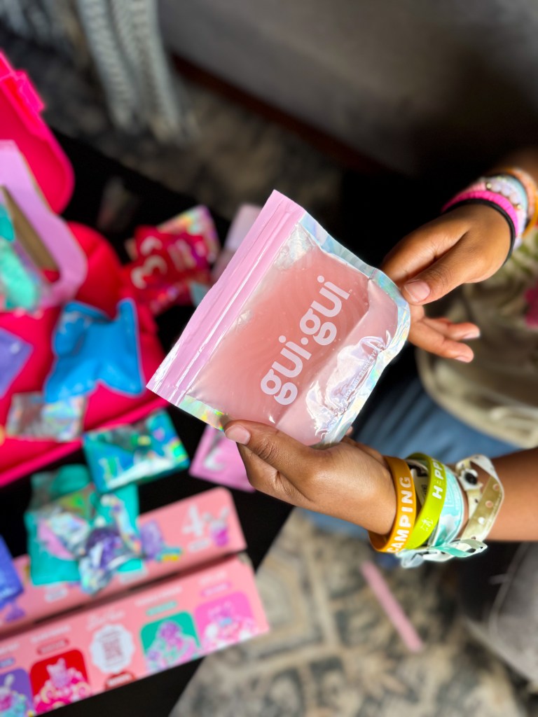A person holding a pink packaging bag labeled 'gui.gui' with colorful accessories and items in the background.