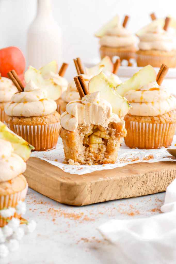 Delicious apple spice cupcakes with creamy frosting, topped with apple slices and cinnamon sticks, displayed on a wooden board.