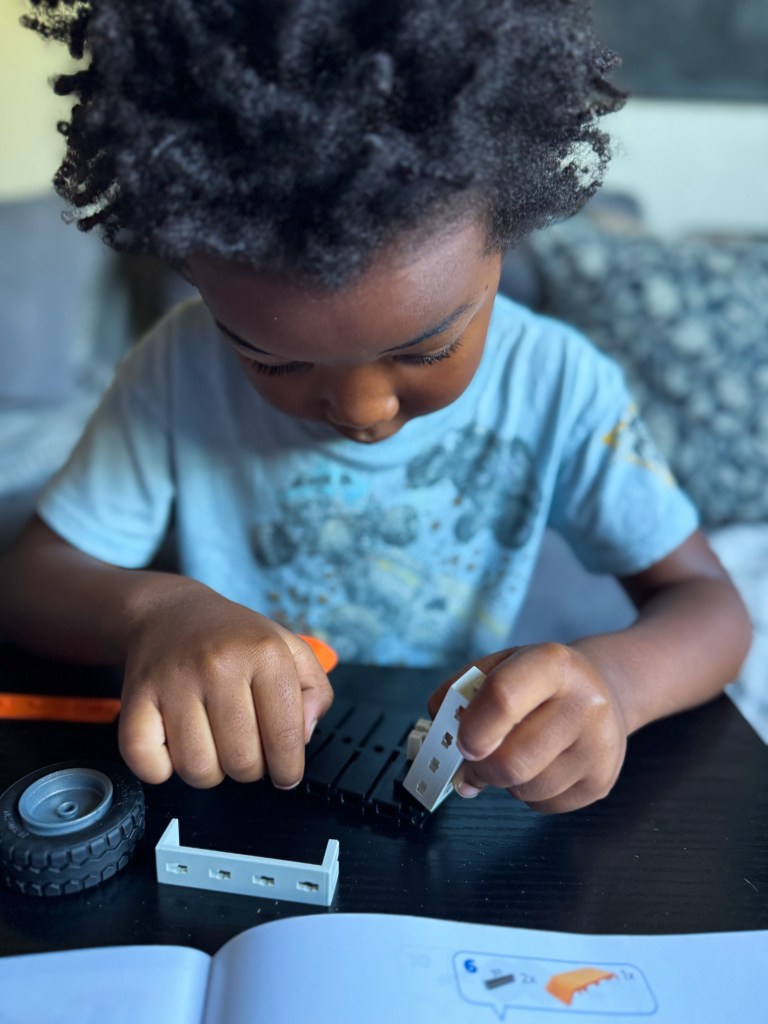 A young child with curly hair focusing on assembling toy parts at a table, with a wheel and tools nearby.