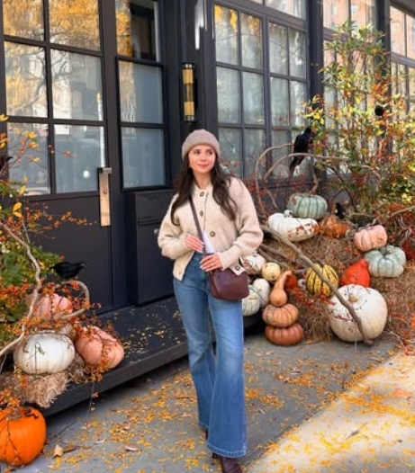 A woman stands in front of a building adorned with pumpkins and autumn foliage, wearing a cozy beige cardigan, wide-leg jeans, and a matching beanie.