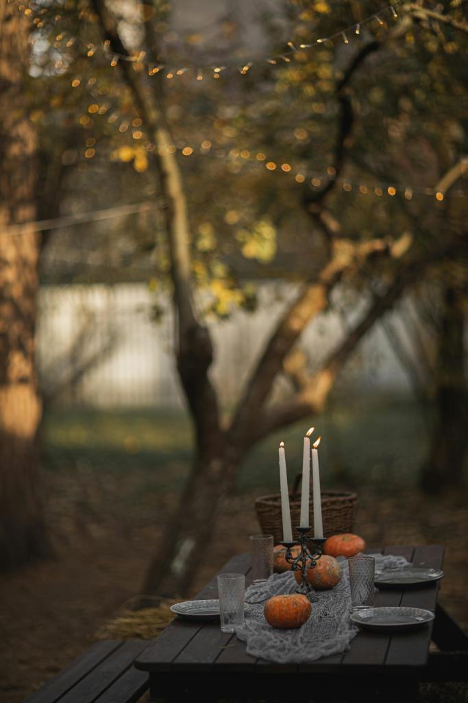 A cozy outdoor table set for a fall gathering, adorned with pumpkins, candles, and string lights, creating a warm and inviting atmosphere.