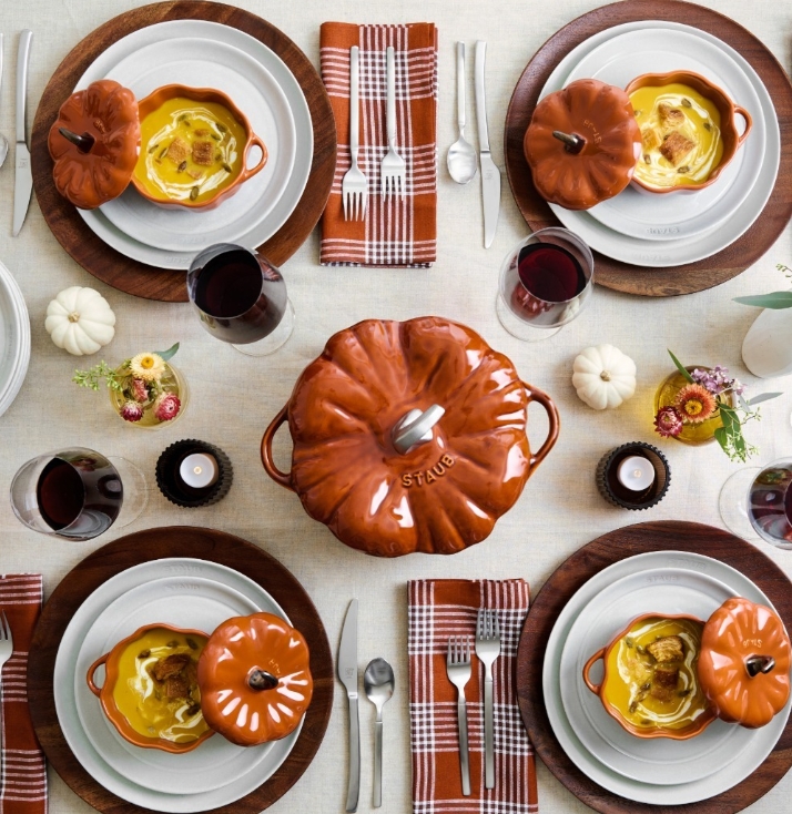 A beautifully styled Thanksgiving table featuring a centerpiece pumpkin-shaped cocotte, white dinnerware, plaid napkins, and rich autumn colors, surrounded by glasses of red wine and small floral arrangements.
