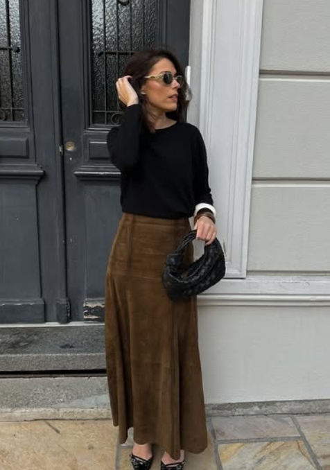 A woman stands in front of a stylish door, wearing a black long-sleeve top paired with a brown suede maxi skirt. She accessorizes with oversized sunglasses and a small black bag, exuding a chic fall look.