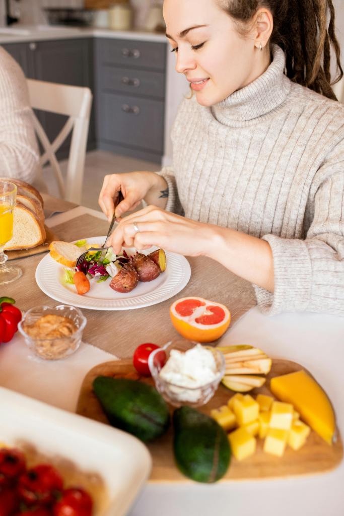 A woman in a beige sweater enjoys a meal at a table, which includes a plate of colorful food and a variety of ingredients like fruits, salad, and cheese.