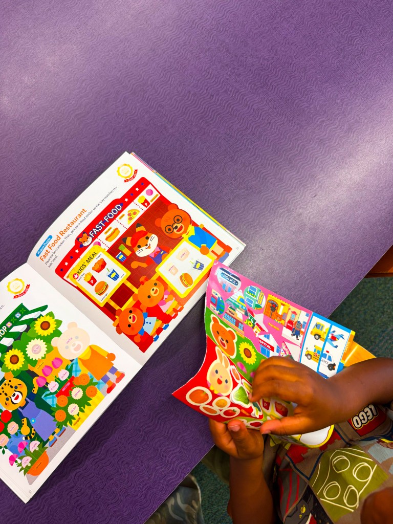A child's hands are holding colorful stickers while a brightly illustrated activity book is open on a purple table, featuring fast food characters and scenes.
