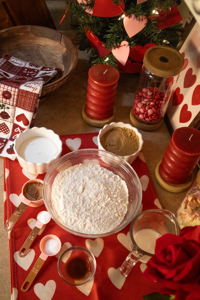 A tabletop scene featuring various baking ingredients including flour, sugar, brown sugar, baking soda, and milk, arranged on a red tablecloth with heart patterns. A red candle, a jar of candies, and a rose are also visible, adding to the festive atmosphere.