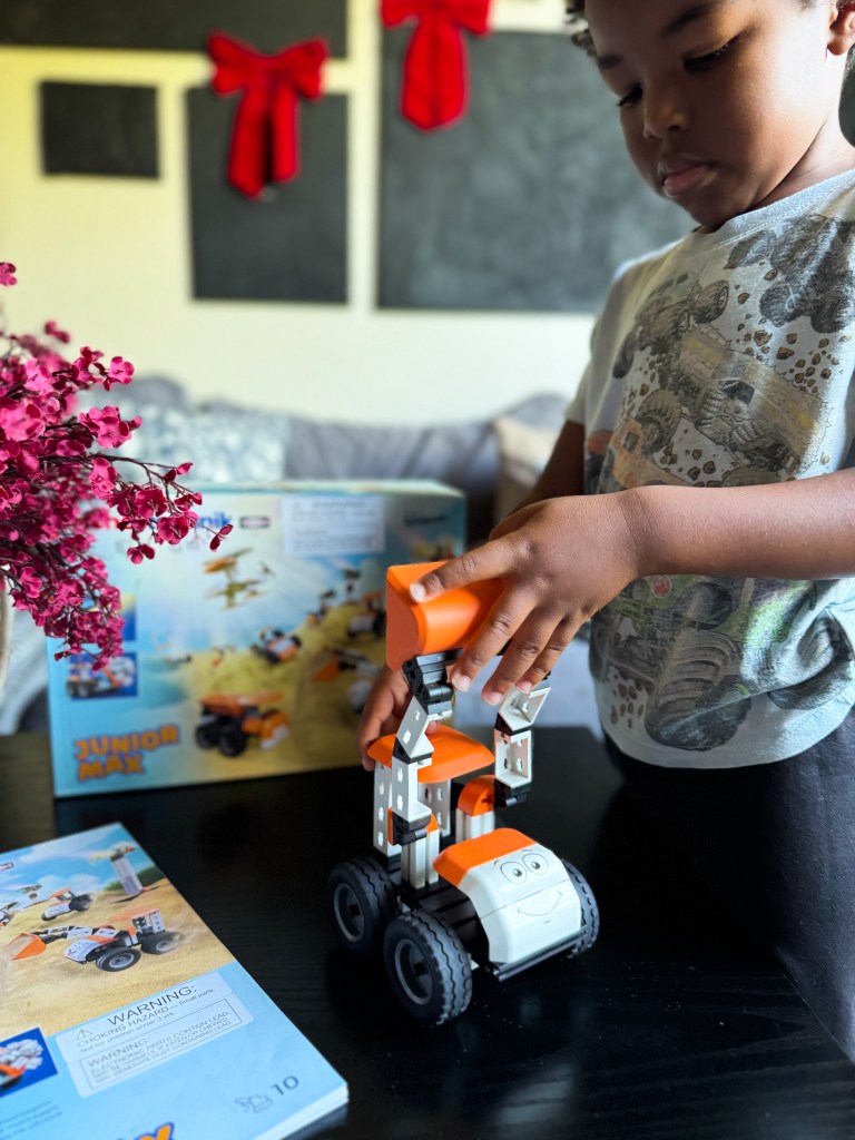 A young child assembling a colorful toy vehicle while sitting at a table with a building instruction manual and a partially completed toy in the background.