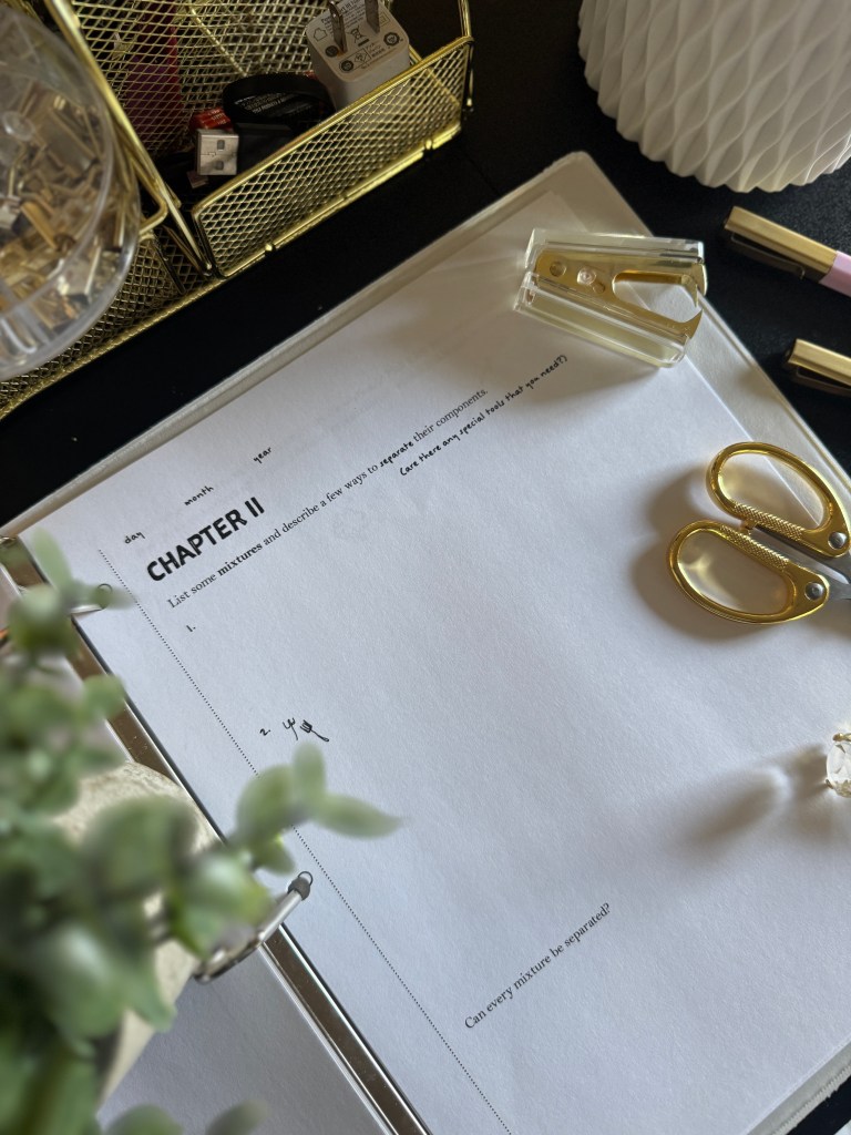 An open notebook with a title page labeled 'CHAPTER II' on a desk, featuring a gold stapler, scissors, and a potted plant in the background.