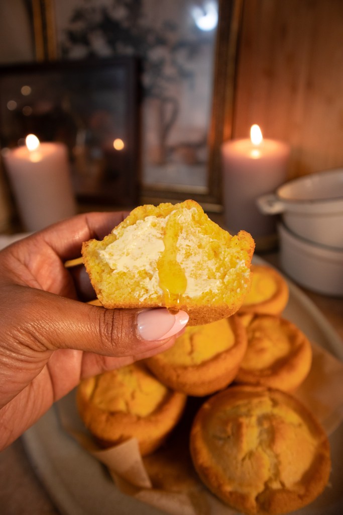 A close-up of a hand holding a half-eaten vegan cornbread muffin filled with a creamy center, with more muffins in the background. Soft candlelight and decorative frames create a warm ambiance.