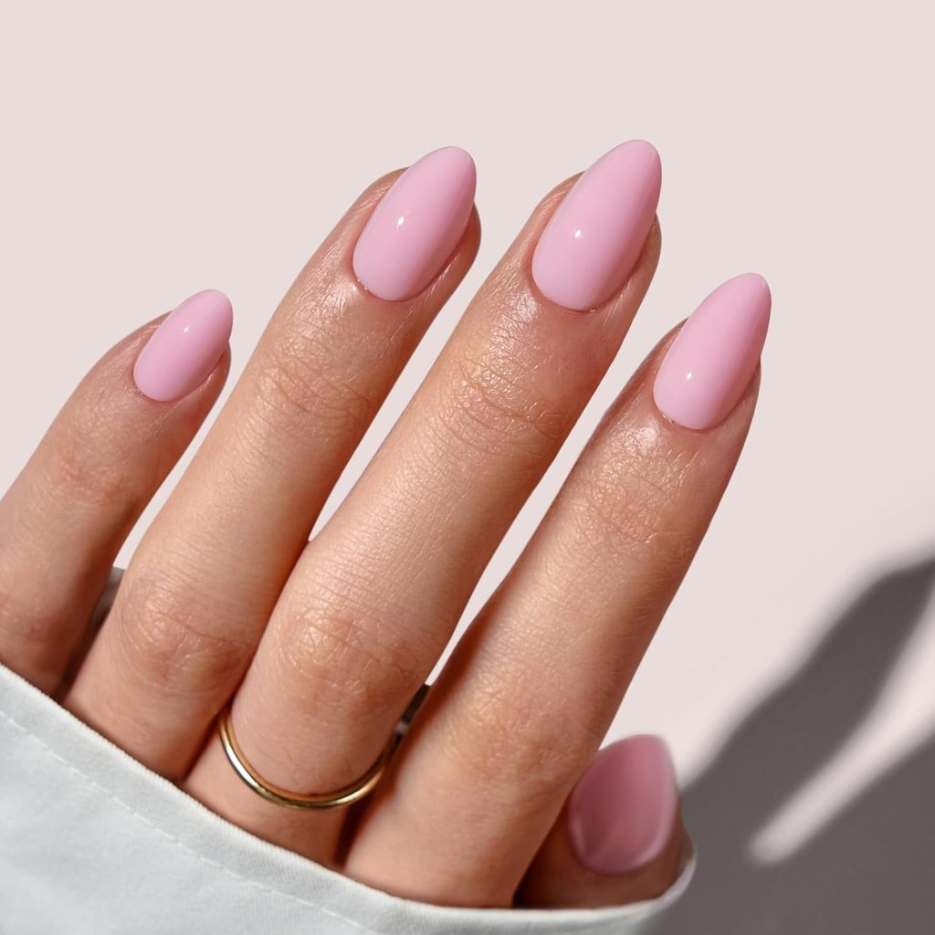 A close-up of a hand with long, almond-shaped nails painted in a soft pink color, adorned with a gold ring on one finger. The hand is partially covered by a light-colored garment.