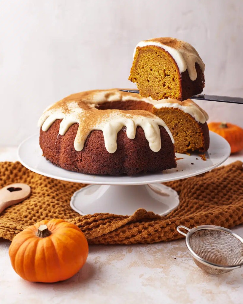 A moist pumpkin bundt cake topped with cream cheese glaze, served on a white cake stand. A small pumpkin and a kitchen tool are nearby, with a cozy backdrop.