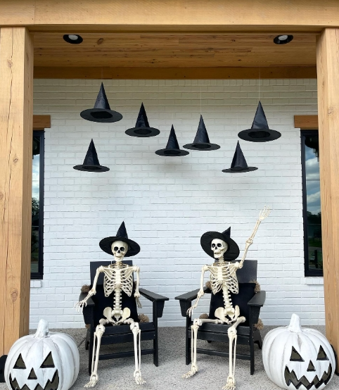 A spooky Halloween porch featuring two skeletons in rocking chairs, each wearing a witch's hat, with hanging black hats above and carved pumpkins on the ground.