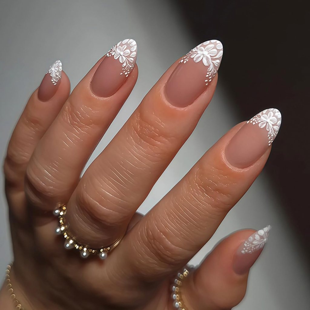 Close-up of a hand showing elegantly manicured nails with a matte finish and white floral designs on the tips. The hand is adorned with several gold rings.