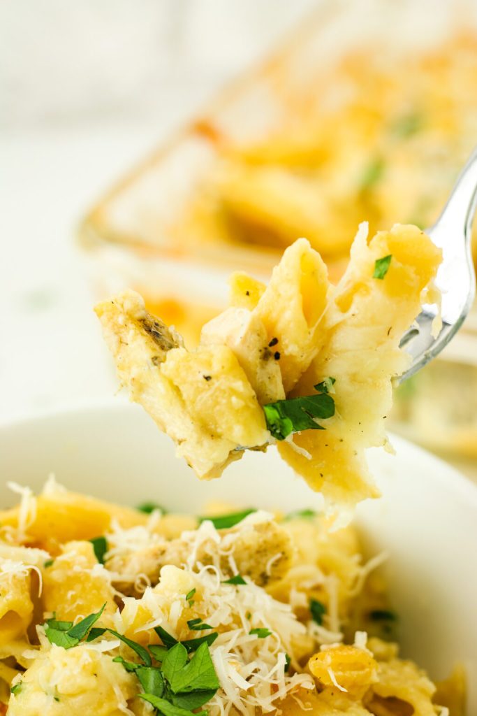 A close-up of a fork lifting a portion of creamy pasta topped with parsley and cheese from a bowl, with a baking dish of pasta in the background.