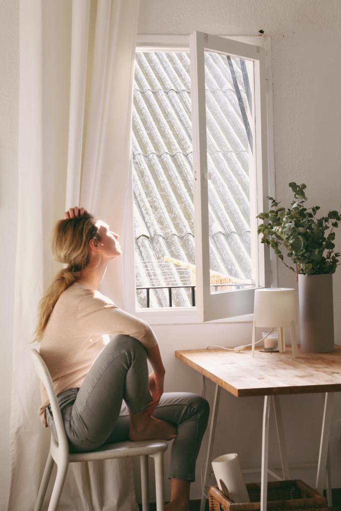 A woman sitting on a chair by an open window, enjoying sunlight while reflecting or relaxing. A potted plant is on the table beside her.