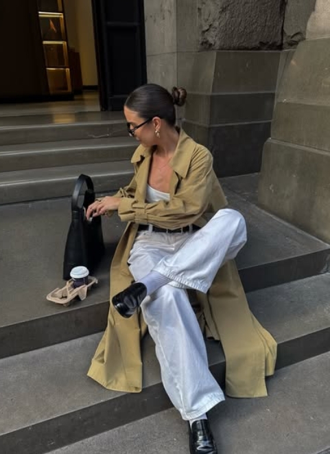 A woman sitting on steps wearing a long beige trench coat, white trousers, and black shoes, holding a small black bag and a coffee cup.