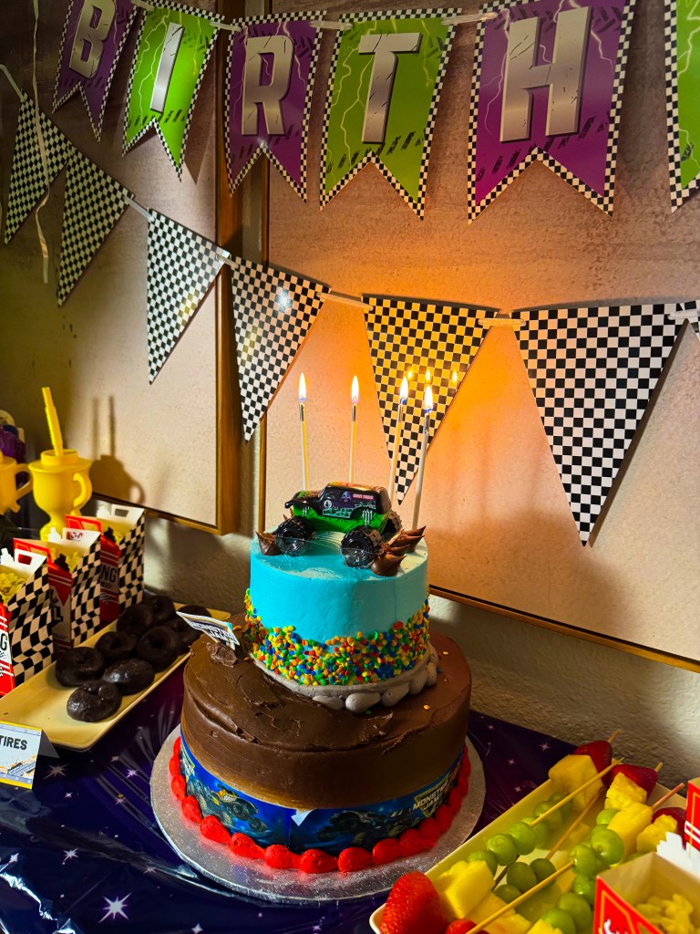 A colorful Monster Jam birthday cake with three layers: chocolate, turquoise, and a layer with sprinkles, topped with a Monster Jam toy truck and lit candles. The background features festive racing  'Birthday' banners and various treats, including fruit and donuts.