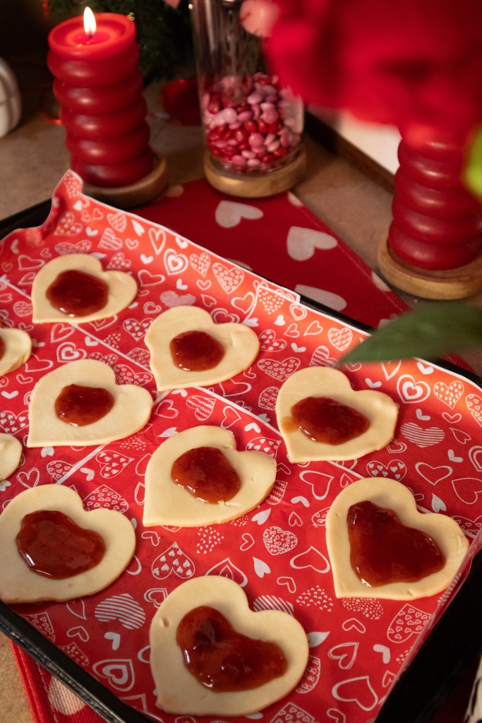 A baking tray with heart-shaped pie crust topped with jelly, set on a red patterned tablecloth surrounded by a lit candle and colorful candies.