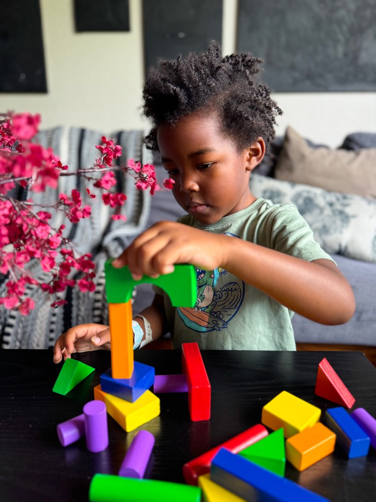 A young child with curly hair is focused on building a structure with colorful blocks on a black table, surrounded by a pink flower arrangement.