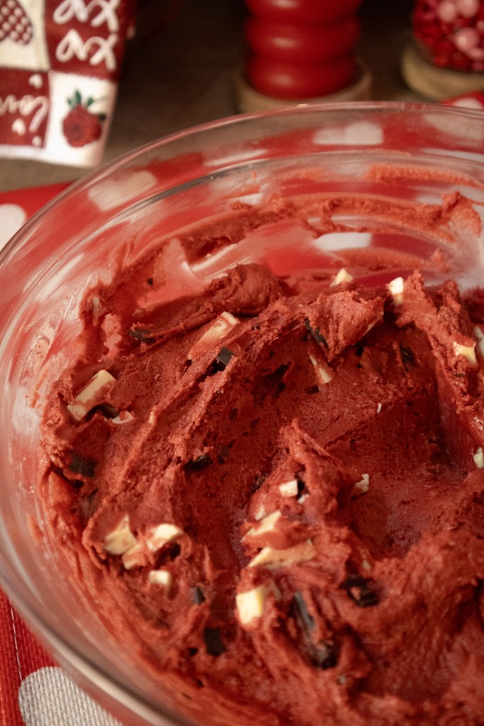 A clear glass bowl filled with rich red velvet cake batter, featuring chunks of white chocolate mixed throughout.