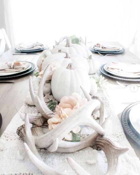 A cozy fall tablescape featuring soft white pumpkins, delicate flowers, and antler accents arranged along a rustic table setting with black plates and linen napkins.