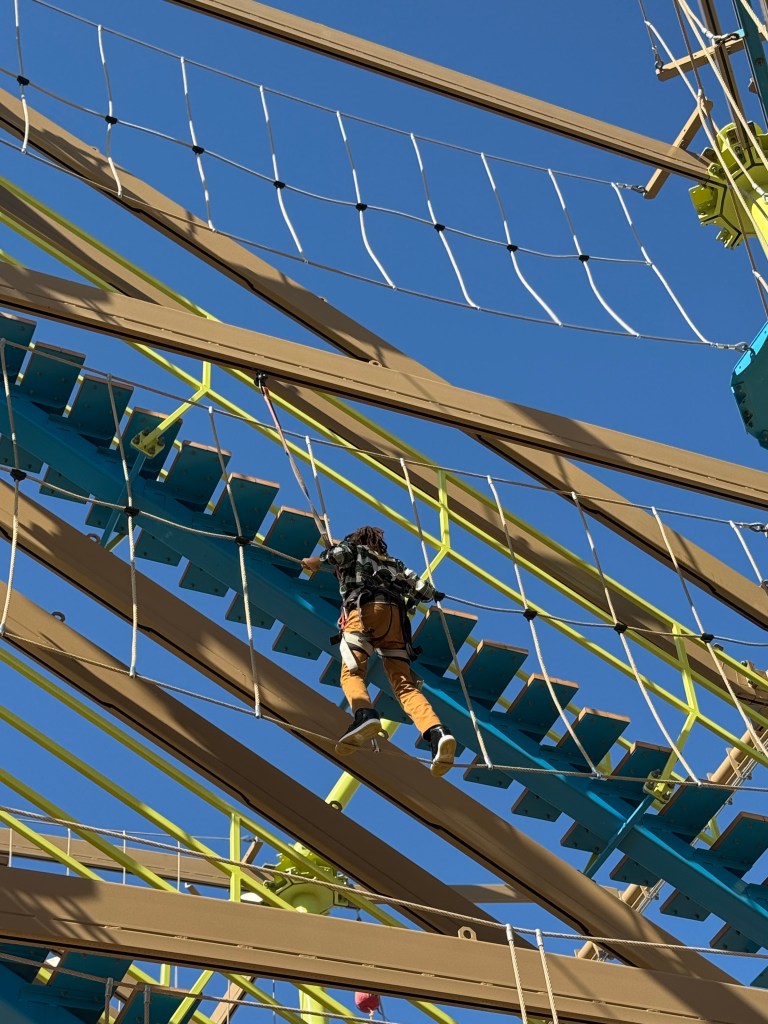 Child crossing a high ropes obstacle course at Great Wolf Lodge Grapevine under a clear blue sky.