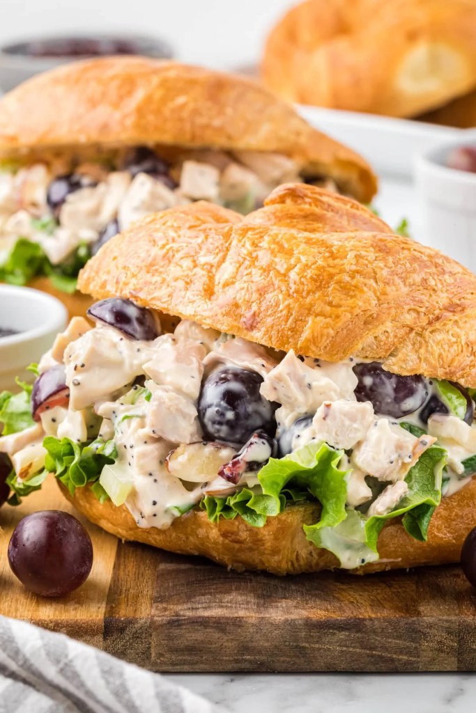 A close-up view of croissant sandwiches filled with chicken salad, grapes, and lettuce, served on a wooden board.