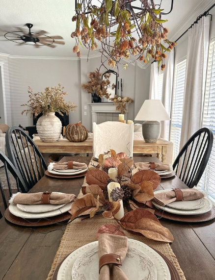 A cozy fall dining table setting featuring beige plates, brown napkins, and dried leaves as a centerpiece, with a rustic wooden table and black chairs.