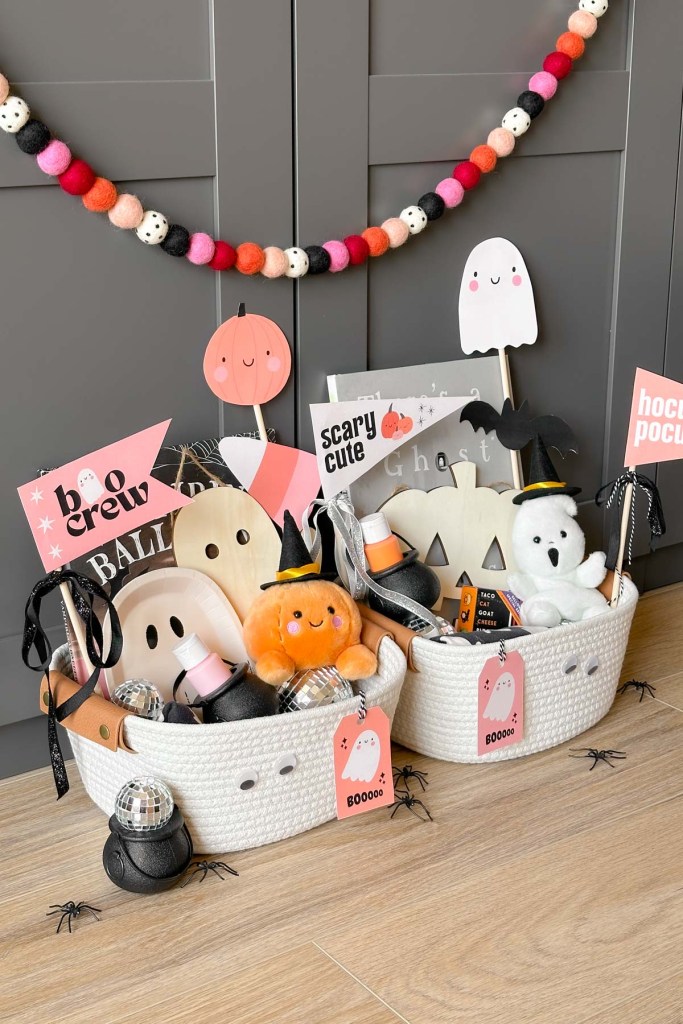 Two woven baskets filled with Halloween-themed goodies including plush toys, decorations, and playful pennants, with a soft pastel color scheme and a festive garland in the background.