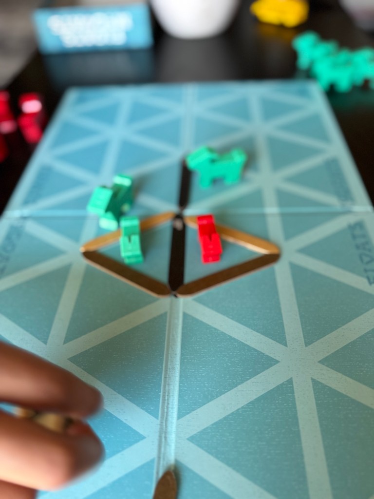A close-up of a board game in progress, featuring colorful game pieces in red and green on a blue playing board. A player's hand is seen in the foreground, poised to make a move.