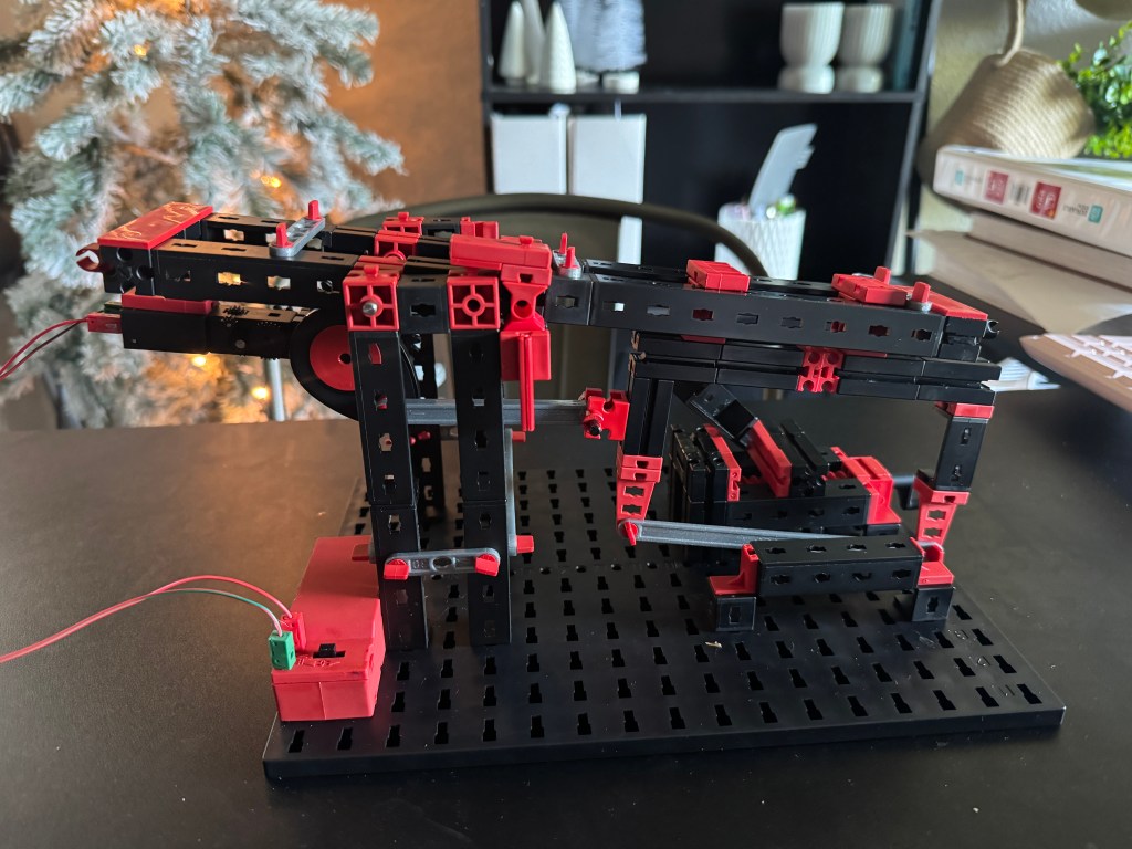 A detailed view of a Fischer Technik STEM kit model, featuring interlocking black and red pieces assembled into a mechanical structure, set on a black base.