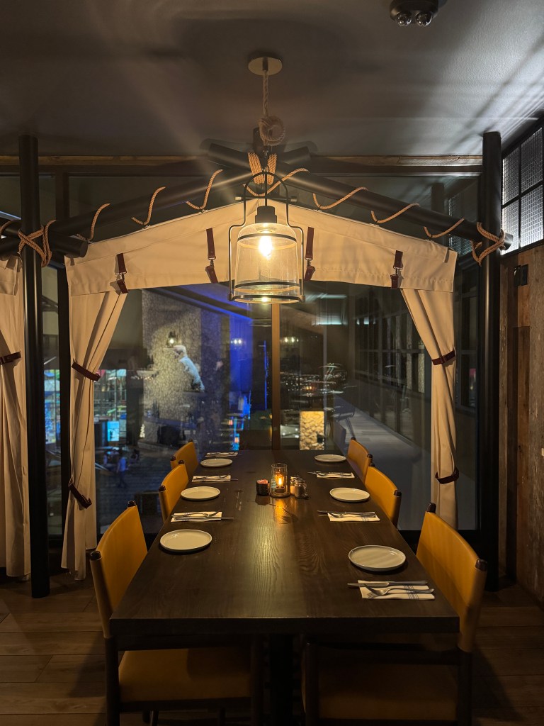 A cozy dining area at Great Wolf Lodge featuring a long wooden table with yellow chairs, set with plates and utensils, illuminated by a hanging lantern, and a view of the indoor water park through large windows.