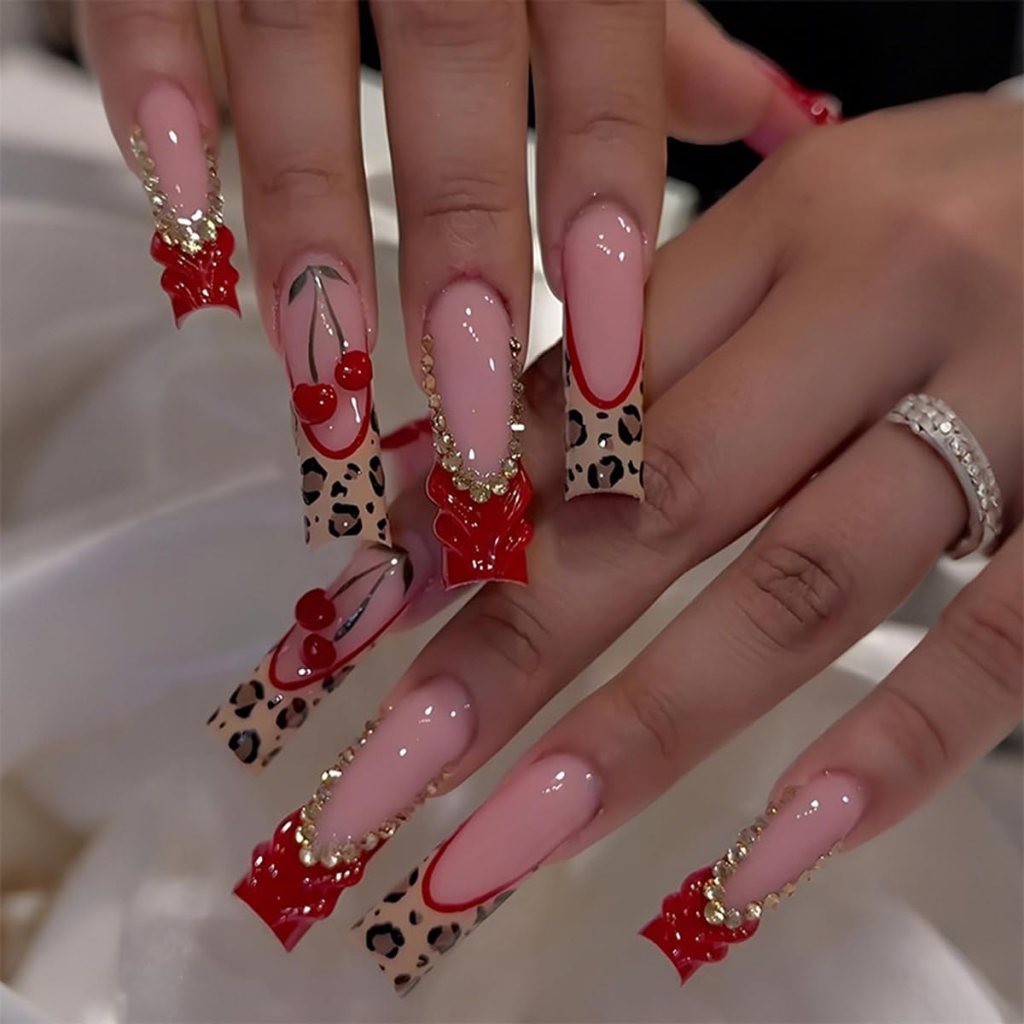 Close-up of manicured hands with long, stylish nails featuring a mix of red and leopard print designs, embellished with gold accents and cherry motifs.