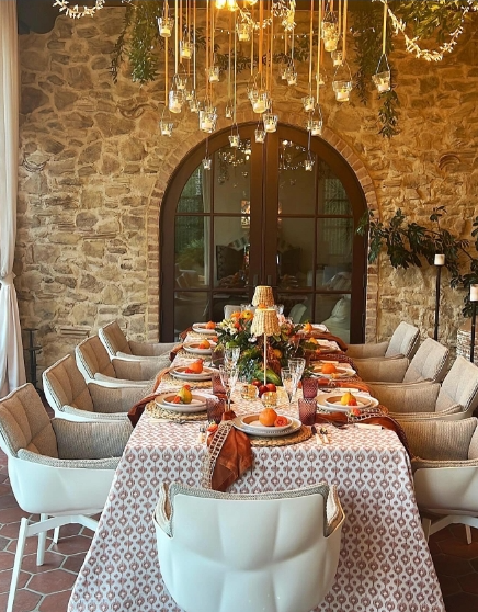 A beautifully set fall dining table featuring a patterned tablecloth, colorful centerpiece of flowers and fruits, and elegant glassware. The setting is surrounded by natural stone walls and hanging lights, creating a warm and inviting atmosphere.